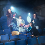 Foto promocional de l’obra La nit dels mons. En una platea de teatre amb butaques blaves i il·luminades per dos focus, quatre persones plantades en mig. A l’esquerra, un home calb amb una jaqueta de pèl marró crida mentre amb un extintor apunta a una dona que es troba en mig de l’escena amb un vestit blanc un llibre a la mà i cara de sorpresa. A la dreta, dues dones amb jaqueta negra i gafes de pasta, criden, una sosté una escombra i un gibrell ple d’aigua que esquitxa l’escena i l’altra a la mà porta la tapa d’un cubell metàl·lic de brossa amb el qual es protegeix.