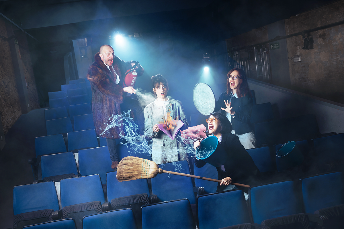Foto promocional de l’obra La nit dels mons. En una platea de teatre amb butaques blaves i il·luminades per dos focus, quatre persones plantades en mig. A l’esquerra, un home calb amb una jaqueta de pèl marró crida mentre amb un extintor apunta a una dona que es troba en mig de l’escena amb un vestit blanc un llibre a la mà i cara de sorpresa. A la dreta, dues dones amb jaqueta negra i gafes de pasta, criden, una sosté una escombra i un gibrell ple d’aigua que esquitxa l’escena i l’altra a la mà porta la tapa d’un cubell metàl·lic de brossa amb el qual es protegeix.