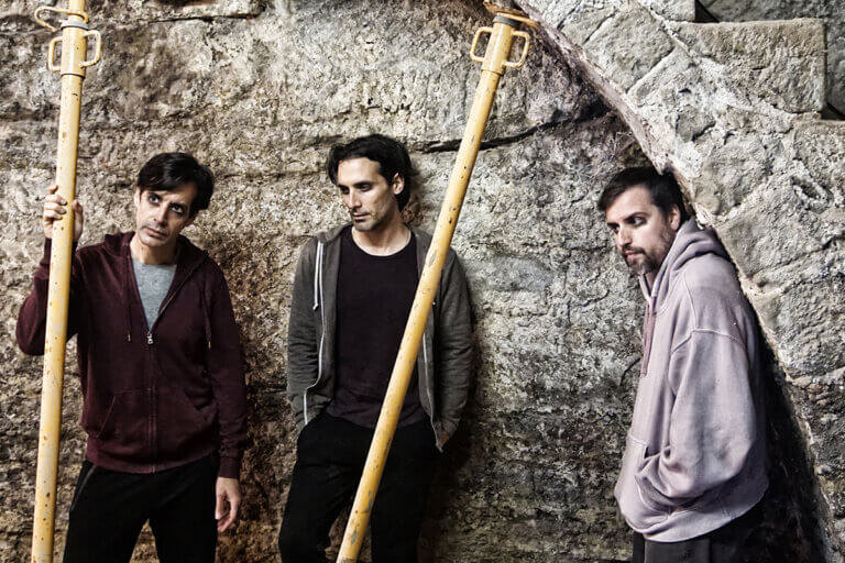 Tres homes vestits de xandalls de colors grisos i bruts de greix dins una volta de pedra suportada per bastides grogues.