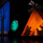 Foto promocional de l’espectacle escolar ‘Agafa el foc i corre!’. A l’esquerra, veiem l’ombra d’un arbre sense fulles, i a la dreta, una tenda de campanya il·luminada de groc. A dins, un grup de persones al voltant del foc: una dona tocant la flauta, un llop assegut al seu costat, una altra dona i dos infants escoltant la música.