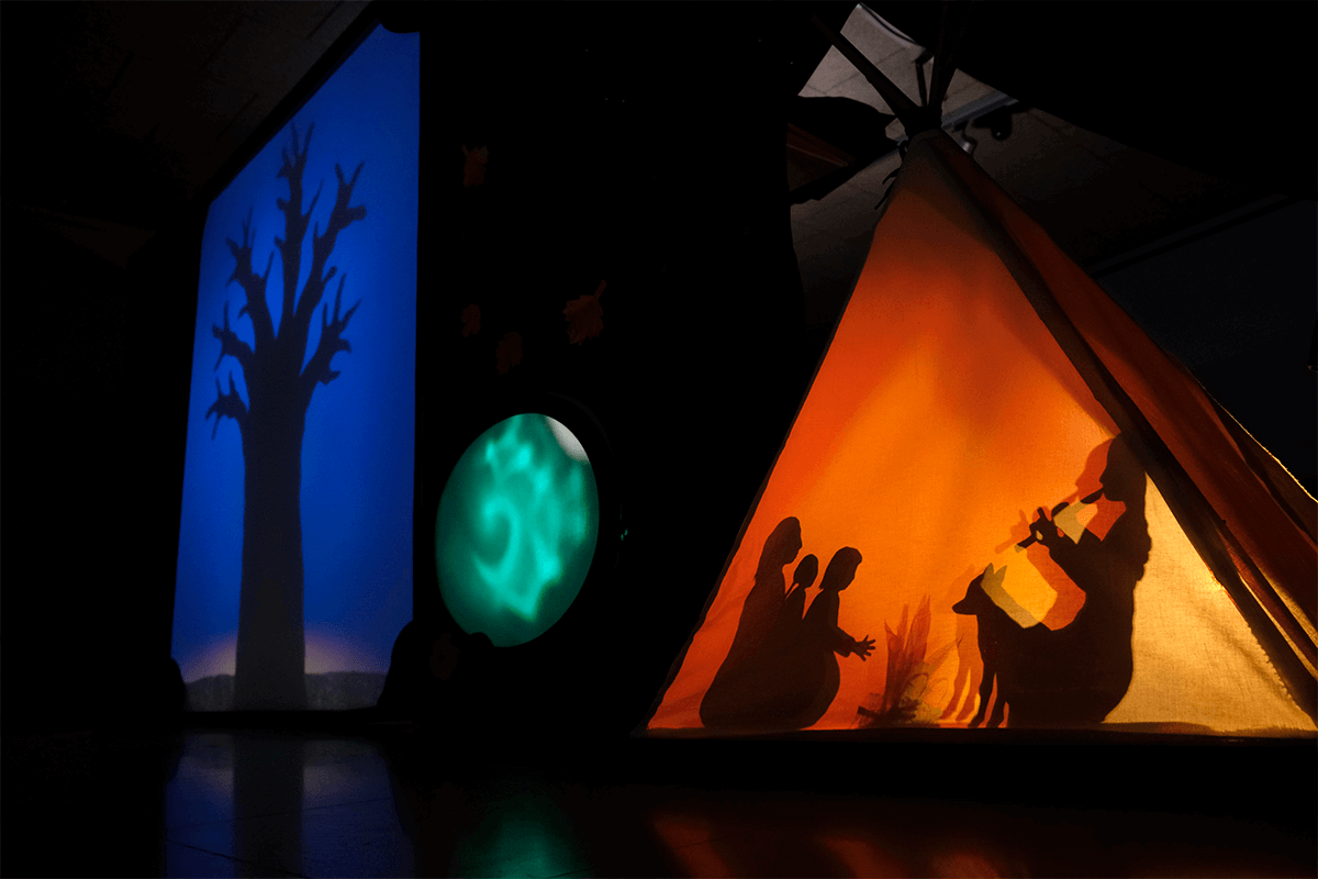 Foto promocional de l’espectacle escolar ‘Agafa el foc i corre!’ A l’esquerra, veiem l’ombra d’un arbre sense fulles, i a la dreta, una tenda de campanya il·luminada de groc. A dins, un grup de persones al voltant del foc: una dona tocant la flauta, un llop assegut al seu costat, una altra dona i dos infants escoltant la música.