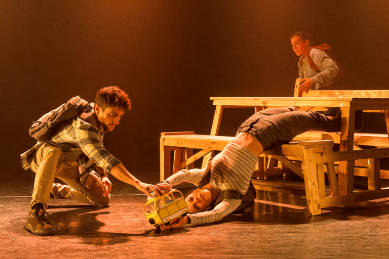 Foto promocional de l’obra Caos. Una llum càlida de tons taronges i grocs il·lumina l’escena. A la dreta, un berenador de fusta del qual sobreïx una dona que de forma expressiva estira el seu cos fins que el seu cap toca terra. Té els braços estirats sostenint una petita caravana de joguines que observa fixament. Un altre home ajupit a terra estén el braç i sosté també la caravana amb la mà. Al fons, darrere del berenador una dona s’apropa, observant l’escena. Totes tres porten motxilles d’excursió a l’esquena.