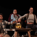 Foto promocional de l’obra Caos. Un home i dues dones vestides amb roba de muntanya i motxilles, assegudes a sobre d’una taula de berenador amb bancs de fusta, totes tres assenyalen una joguina en forma d’autobús groc que es troba al mig de la taula.