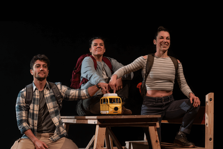 Foto promocional de l’obra Caos. Un home i dues dones vestides amb roba de muntanya i motxilles, assegudes a sobre d’una taula de berenador amb bancs de fusta, totes tres assenyalen una joguina en forma d’autobús groc que es troba al mig de la taula.