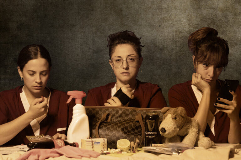 Foto promocional de l'obra Com eliminar l'olor de lleixiu de les mans. En una imatge en tons sèpia amb un fons gris, hi ha en primer pla una taula on de forma desorganitzada es disposa una bossa de marca de cuir marró, un peluix de gos, mitja llima, un pot de neteja, uns guants de plàstic rosa, pastilles, una cervesa, unes llaminadures, tabac d'embolicar i una botella de Coca-Cola. Assegudes hi ha tres dones vestides amb bates granats iguals, una mira el mòbil, l'altra amb un braç embenat mira fixament a càmera i la tercera mira fixament una llaminadura.