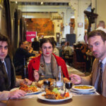 Foto promocional de l’obra Els carnissers. En un restaurant d’un celler de vins atapeït de gent, tres persones mengen un menú casolà de pollastre amb patates, a l’esquerra un home amb jaqueta de vellut marró i un boà de pèl marró, al centre una dona amb una jaqueta vermella combinada amb els llavis i els cabells recollits, i a l’esquerra un home ros amb una jaqueta marró i una corbata blava, tots tres miren al davant fixament en silenci.