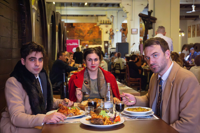 Foto promocional de l’obra Els carnissers. En un restaurant d’un celler de vins atapeït de gent, tres persones mengen un menú casolà de pollastre amb patates, a l’esquerra un home amb jaqueta de vellut marró i un boà de pèl marró, al centre una dona amb una jaqueta vermella combinada amb els llavis i els cabells recollits, i a l’esquerra un home ros amb una jaqueta marró i una corbata blava, tots tres miren al davant fixament en silenci.