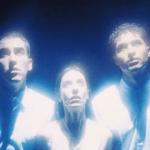 Foto promocional de l’obra ‘Goodbye Europe. Lost words’. Tres intèrprets, dos hòmens i una dona miren fixament cap a dalt. Observen amb molta atenció alguna cosa que l’espectador no pot veure. Tots tres van vestits amb camises blanques corporatives. La llum de l’escena és blava i el fons d’un blau marí intens. De les seves figures surten feixos de llum blanca i blava.