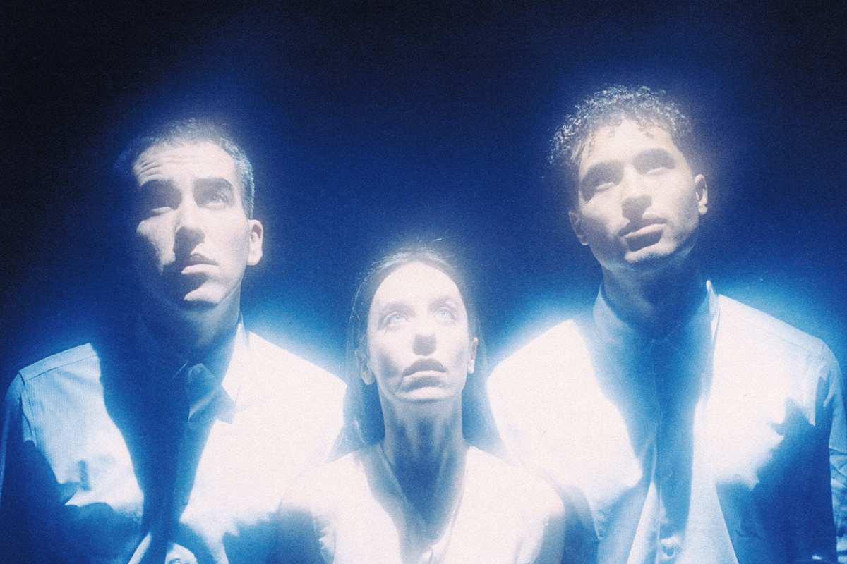 Foto promocional de l’obra ‘Goodbye Europe. Lost words’. Tres intèrprets, dos hòmens i una dona miren fixament cap a dalt. Observen amb molta atenció alguna cosa que l’espectador no pot veure. Tots tres van vestits amb camises blanques corporatives. La llum de l’escena és blava i el fons d’un blau marí intens. De les seves figures surten feixos de llum blanca i blava.