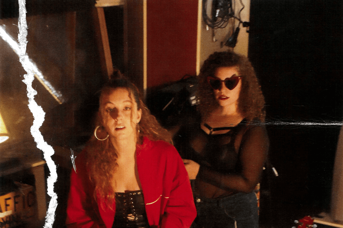 Foto promocional de l'obra Kathleen Hannah. Dues dones mirant cap a la càmera, una al davant amb jaqueta vermella i arracades grans, i una altra darrere amb ulleres de sol en forma de cor. La imatge té ratllades i una gran marca blanca al costat esquerre.