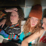 Foto promocional de Kathleen Hanna. En primer pla, tres dones joves posen per a una foto. La dona de l’esquerra s’aparta els cabells de la cara i mira directament a càmera. Va vestida amb una dessuadora amb un estampat dels noranta, porta una guitarra al damunt folrada amb adhesius de colors. La dona a la seva dreta li passa el braç per les espatlles i també mira directament a càmera. Porta un barret de llana i una camisa de quadres. A la dreta del tot, una dona amb els cabells negres recollits i una samarreta blanca de tirants amb unes lletres vermelles mira les seves companyes.