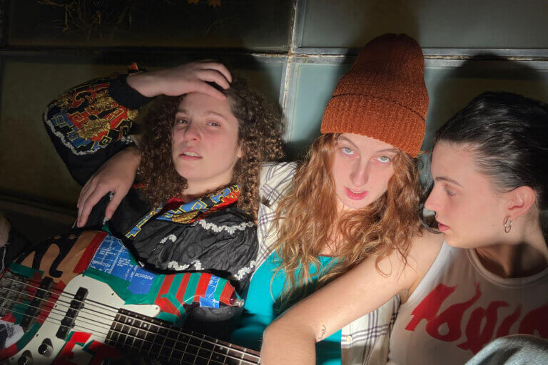 Foto promocional de Kathleen Hanna. En primer pla, tres dones joves posen per a una foto. La dona de l’esquerra s’aparta els cabells de la cara i mira directament a càmera. Va vestida amb una dessuadora amb un estampat dels noranta, porta una guitarra al damunt folrada amb adhesius de colors. La dona a la seva dreta li passa el braç per les espatlles i també mira directament a càmera. Porta un barret de llana i una camisa de quadres. A la dreta del tot, una dona amb els cabells negres recollits i una samarreta blanca de tirants amb unes lletres vermelles mira les seves companyes.