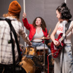 Foto promocional de l'obra Kathleen Hannah. En una sala de fusta amb una catifa, en mig una dona de cabells llargs morenos i arrissats amb una dessuadora vermella toca una bateria, en primer pla a l'esquerra s’intueix una persona amb camisa de quadros color marró i una guitarra, a la dreta hi ha una dona morena que porta una cua, una samarreta blanca de tirants amb lletres vermelles formant flames i una guitarra elèctrica, que mira cap a la bateria.