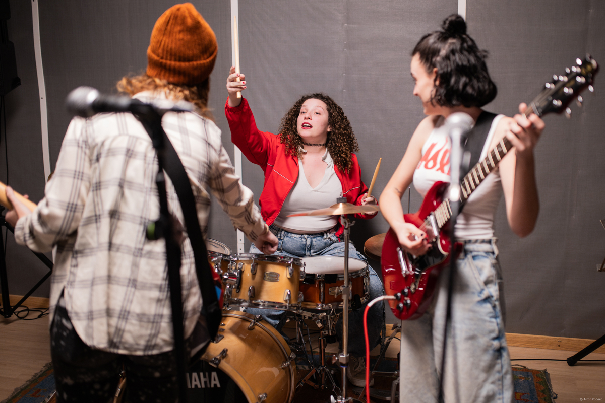 Foto promocional de l'obra Kathleen Hannah. En una sala de fusta amb una catifa, en mig una dona de cabells llargs morenos i arrissats amb una dessuadora vermella toca una bateria, en primer pla a l'esquerra s’intueix una persona amb camisa de quadros color marró i una guitarra, a la dreta hi ha una dona morena que porta una cua, una samarreta blanca de tirants amb lletres vermelles formant flames i una guitarra elèctrica, que mira cap a la bateria.