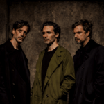 Foto promocional de ‘Les mans brutes’.Tres hòmens drets sobre un fons obscur que sembla de pedra bruta. Dos d’ells, els situats a la dreta i el mig, romanen de perfil orientats cap a l’esquerra. El de l’esquerra va vestit de negre i el del mig amb una gavardina verda llarga. Tots dos miren a un punt indefinit fora de pla amb actitud seriosa i pensativa. El tercer home, situat a l’esquerra de la imatge i vestit de negre, mira a un punt indefinit darrere de l'objectiu de la càmera. Tots tres semblen tenir als ulls una ombra negra.