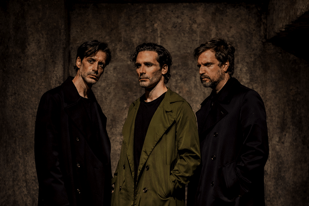 Foto promocional de ‘Les mans brutes’.Tres hòmens drets sobre un fons obscur que sembla de pedra bruta. Dos d’ells, els situats a la dreta i el mig, romanen de perfil orientats cap a l’esquerra. El de l’esquerra va vestit de negre i el del mig amb una gavardina verda llarga. Tots dos miren a un punt indefinit fora de pla amb actitud seriosa i pensativa. El tercer home, situat a l’esquerra de la imatge i vestit de negre, mira a un punt indefinit darrere de l'objectiu de la càmera. Tots tres semblen tenir als ulls una ombra negra.