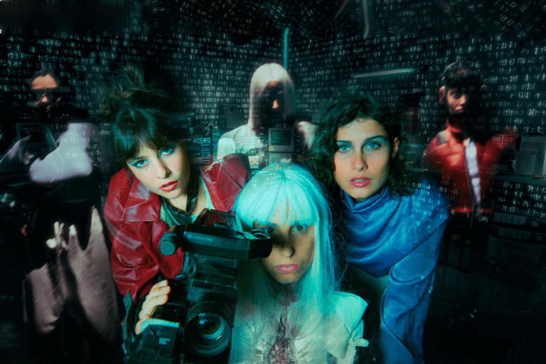Foto promocional Mary’s Room black box. Sobre un fons fosc tres dones en primer pla. Dues al darrere, una amb cabells foscos, curt i una cua alta, vestida amb una jaqueta de cuir vermell, l’altra amb cabells foscos curts i arrissats amb una camisa blava metàl·lica, i al front, una dona amb els cabells blancs, llisos i llargs i un mono de cuir sintètic de color negre amb franges blanques amb una càmera de vídeo.
