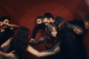 Foto promocional de ‘Salomé’. Sis intèrprets vestits de negre sobre un fons vermell fosc. Tots es troben en moviment, s’entrellacen, s’agafen i s’estiren. La imatge té uns feixos de llum que imiten la sensació d’un moviment ràpid i, al centre de la fotografia, una dona jove intenta escapolir-se de les mans que tracten de retenir-la agafant-la per les costelles. La seva expressió és d’esforç i a sobre d’ella els feixos de llum són més intensos.