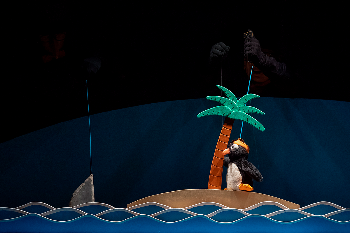 Un titellaire manipula un pingüí que es troba a la vora d'una illa de palmeres, mentre un altre titellaire manipula un tauró, veiem l'aleta sobresortir de l'aigua.