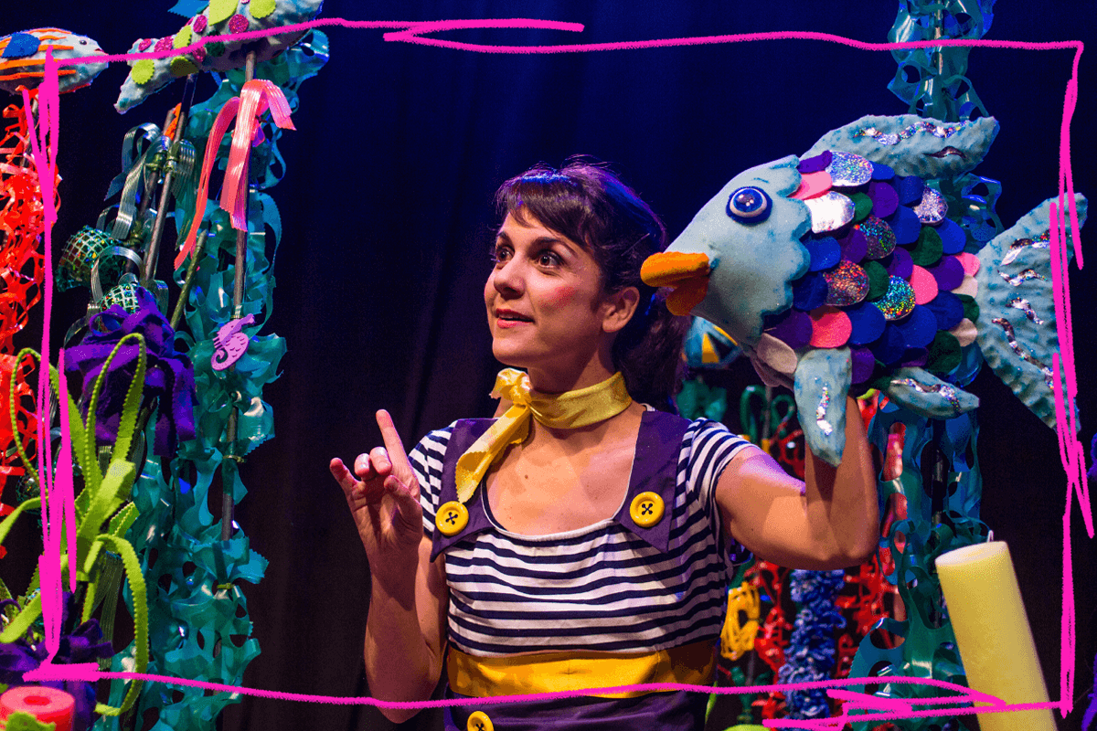 Foto promocional de l’espectacle escolar ‘El peix irisat’. Una dona amb un vestit de marinera i uns botons enormes de color groc subjecta una titella d’un peix amb escates de color blau, lila, rosat i escates brillants. Al voltant, algues marines multicolors i molts peixos.