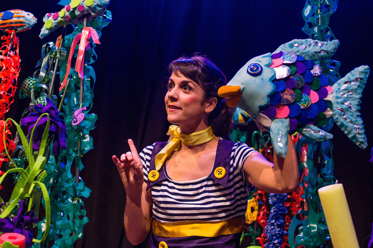 Foto promocional de l’espectacle escolar ‘El peix irisat’. Una dona amb un vestit de marinera i uns botons enormes de color groc subjecta una titella d’un peix amb escates de color blau, lila, rosat i escates brillants. Al voltant, algues marines multicolors i molts peixos.