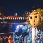 Fotografia promocional de l'obra FILLKE PEWMA (Sueños). A l’aire lliure i de nit, en primer pla a la dreta, una persona amb una màscara de fusta i un vestit recobert de cintes de colors metàl·lics i lluents en tons roses, violetes, blaus i platejats mira a càmera enfocant a l’esquerra de l’objectiu amb una llanterna de llum blanca. La seva mà està coberta per un guant blanc. Darrere seu una barana de ferro, que el separa d’un riu d’aigües fosques i un pont il·luminat per llums de de tons càlids. Sembla el paisatge d’una ciutat.