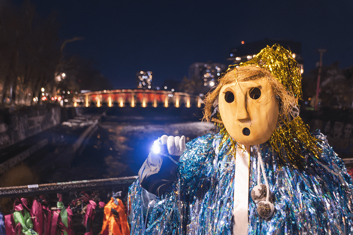 Fotografia promocional de l'obra FILLKE PEWMA (Sueños). A l’aire lliure i de nit, en primer pla a la dreta, una persona amb una màscara de fusta i un vestit recobert de cintes de colors metàl·lics i lluents en tons roses, violetes, blaus i platejats mira a càmera enfocant a l’esquerra de l’objectiu amb una llanterna de llum blanca. La seva mà està coberta per un guant blanc. Darrere seu una barana de ferro, que el separa d’un riu d’aigües fosques i un pont il·luminat per llums de de tons càlids. Sembla el paisatge d’una ciutat.
