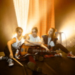 Foto promocional de l’espectacle “Hora Bruixa”. Tres dones assegudes a terra sobre un fons del qual surten feixos de llum daurada. Les tres intèrprets es troben una al costat de l’altra i porten diferents elements musicals. La dona de l’esquerra duu unes baquetes a la mà, la del mig porta una guitarra recolzada sobre la seva cama i la de l’esquerra porta un micro a la mà. Les dues primeres dones miren a càmera desafiants mentre que la tercera mira a les seves companyes. Totes tres van vestides amb faldilles a quadres de diferents colors, samarretes blanques amb iconografia punk i sabatilles Convers. Al costat d’elles podem veure un titella tirat a terra i al fons un teló de quadres escocesos.