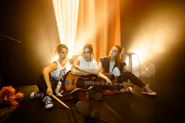 Foto promocional de l’espectacle “Hora Bruixa”. Tres dones assegudes a terra sobre un fons del qual surten feixos de llum daurada. Les tres intèrprets es troben una al costat de l’altra i porten diferents elements musicals. La dona de l’esquerra duu unes baquetes a la mà, la del mig porta una guitarra recolzada sobre la seva cama i la de l’esquerra porta un micro a la mà. Les dues primeres dones miren a càmera desafiants mentre que la tercera mira a les seves companyes. Totes tres van vestides amb faldilles a quadres de diferents colors, samarretes blanques amb iconografia punk i sabatilles Convers. Al costat d’elles podem veure un titella tirat a terra i al fons un teló de quadres escocesos.