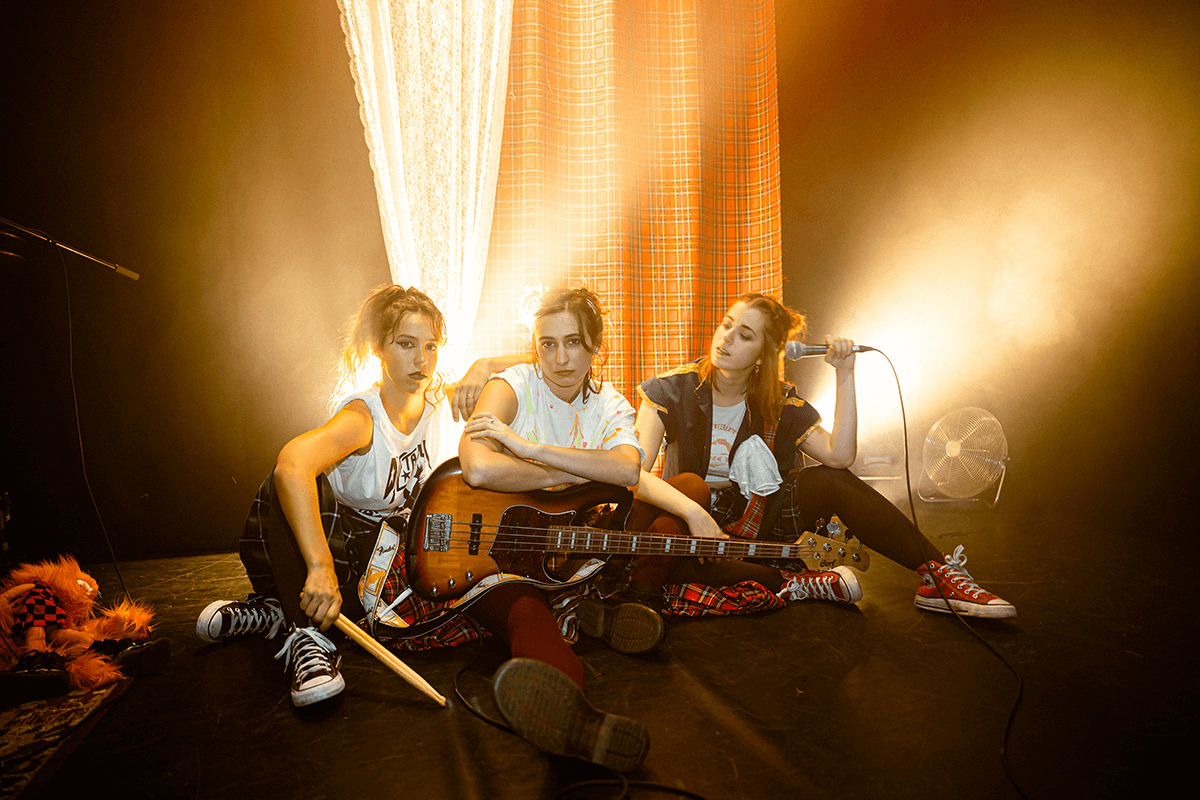 Foto promocional de l’espectacle “Hora Bruixa”. Tres dones assegudes a terra sobre un fons del qual surten feixos de llum daurada. Les tres intèrprets es troben una al costat de l’altra i porten diferents elements musicals. La dona de l’esquerra duu unes baquetes a la mà, la del mig porta una guitarra recolzada sobre la seva cama i la de l’esquerra porta un micro a la mà. Les dues primeres dones miren a càmera desafiants mentre que la tercera mira a les seves companyes. Totes tres van vestides amb faldilles a quadres de diferents colors, samarretes blanques amb iconografia punk i sabatilles Convers. Al costat d’elles podem veure un titella tirat a terra i al fons un teló de quadres escocesos.