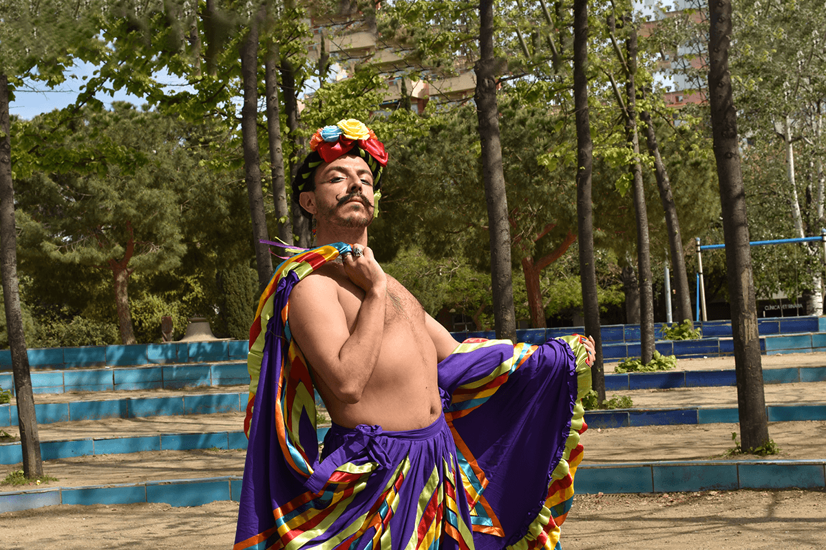 Foto promocional de l’espectacle “Quería ser maricón pero nací sudaca”. A la imatge apareix l'artista Mazorra mirant a càmera desafiant amb la barbeta alta. Va vestida amb unes faldilles de colors morats, rojos, grocs i blaus que agafa pels extrems amb les mans. L'extrem de l'esquerra el sosté en la mà que reposa sobre el seu muscle generant com una mena de capa. Porta el pit nu i unes flors de colors al cap. També porta els ulls pintats i un bigoti i barba pentinats.