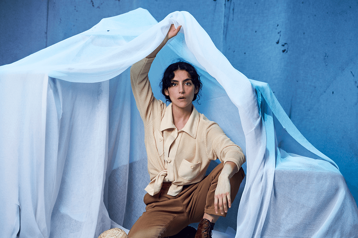 Fotografia promocional de l'obra Mi madre nada. Una actriu amb barret de palla, camisa beix i pantalons de color caqui salta amb una cama doblegada mentre s'aguanta el barret amb una mà i arrossega amb l'altra una tela blanca. Porta els cabells recollit i té expressió de sorpresa. Al fons, una paret blau clar amb les paraules escrites a mà "26 de agosto 2020" i una lletra "a" en cursiva.