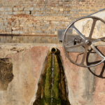 Primer pla d'una font de pedra antiga: un broc central deixa caure un petit raig d'aigua sobre una superfície humida amb molsa verda. A la dreta, una roda metàl·lica gran forma part del mecanisme de la font. Al fons, un mur de pedra irregular amb restes d'un arc de maó suggereix un entorn històric o patrimonial.