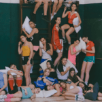 Un grup nombrós de joves vestits amb roba esportiva i coixins posen en un gimnàs escolar amb escales de fusta verdes. Alguns estan drets, d'altres asseguts i d'altres estirats a terra, en una composició piramidal que transmet cansament i complicitat.