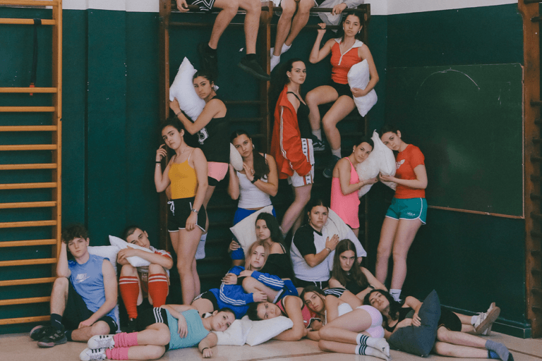 Un grup nombrós de joves vestits amb roba esportiva i coixins posen en un gimnàs escolar amb escales de fusta verdes. Alguns estan drets, d'altres asseguts i d'altres estirats a terra, en una composició piramidal que transmet cansament i complicitat.