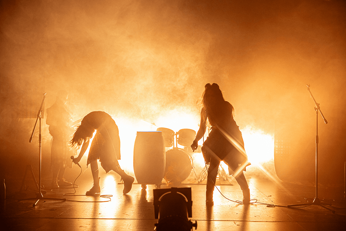 Foto promocional d’Hora Bruixa. Siluetes de dos actrius contra una intensa llum daurada i fum dens en un escenari de rock. Una s'inclina cap al terra amb un micròfon de forma expressiva, mentre l’altra arrossega els cables d’un micro. L'atmosfera és potent i onírica.